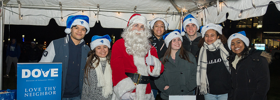 Toy recipients in a warehouse holding up a sign saying "I Love Seton Hall" and wearing santa hats. There are wrapped gifts at their feet for children. 