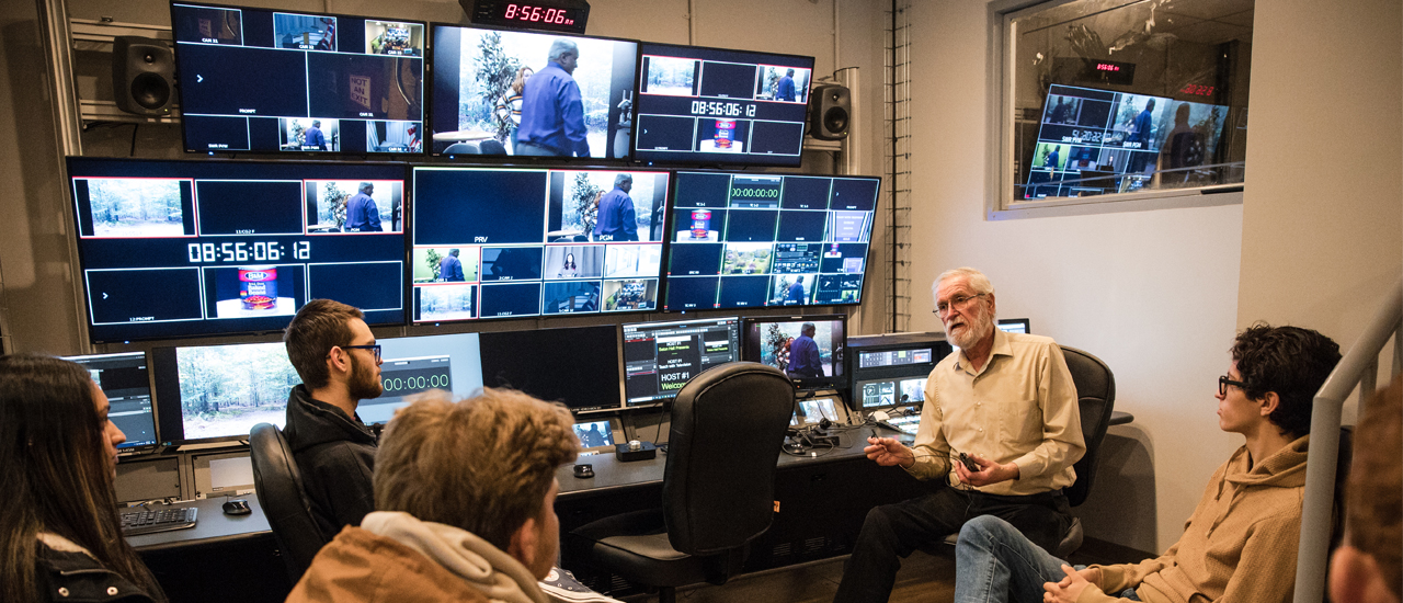 Image of a professor and five studios in a video and audio studio with several monitors behind them showing a live recording happening in another room. 