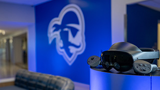 Virtual reality headset and controllers displayed in front of a large Seton Hall Pirate logo on a blue wall.